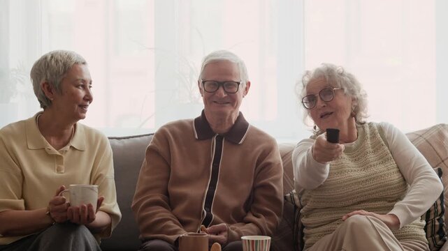 Stab POV shot of three elderly adults switching channels with remote controller while sitting on sofa in front of TV gathering at home