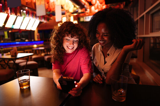 Diverse friends laughing, connecting, viewing content on smartphone at bar