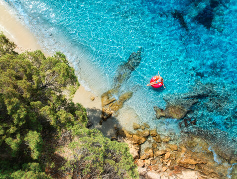 Aerial drone view of woman on pink inflatable ring in turquoise water near rocky coast and sandy beach with green trees. Summer in Mallorca, Balearic Islands, Spain. Mediterranean sea. Top down view