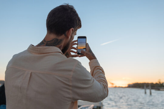 Man photographing sunset with smartphone by the water