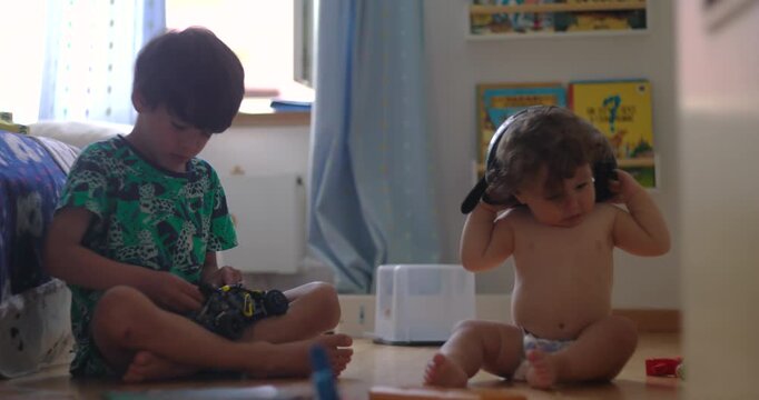 Two children sitting on floor playing together while baby tries to wear oversized helmet, older boy focused on toy car, authentic sibling interaction and playful home moment