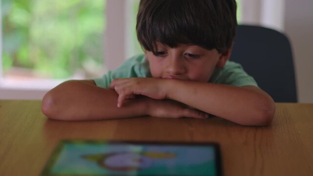Young boy leaning on table watching tablet with distant expression showing quiet absorption low engagement and reflective mood during calm screen time moment at home