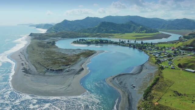 A boat speeds down the turquoise waters of the River in Marokopa, Waikato, New Zealand, passing by lush green hills and a small coastal town.