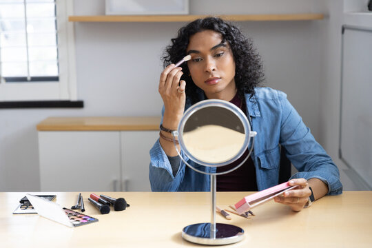 Non-binary person wearing denim shirt applying eyeshadow with brush at light wood table with mirror