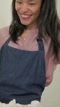 Vertical video: Leaning baker, apron, pink headband reaching kitchen counter, sprinkling cupcakes