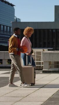 Vertical video: Entering woman pulling suitcase, man walking holding basketball across plaza