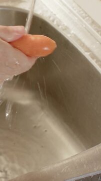 Vertical video: Running faucet, woman's hands washing carrot at kitchen sink, preparing for cooking