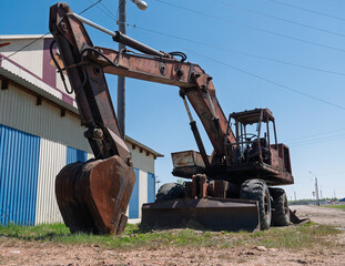 Weathered wheeled excavator stands idle near a striped building on a sunny day © Evgenii Bakhchev