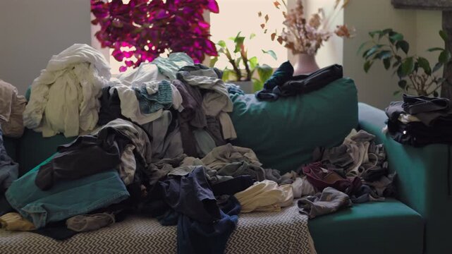 Camera slowly reveals clothes pile extending across sofa with additional garments scattered nearby emphasizing accumulation and domestic workload in lived in space
