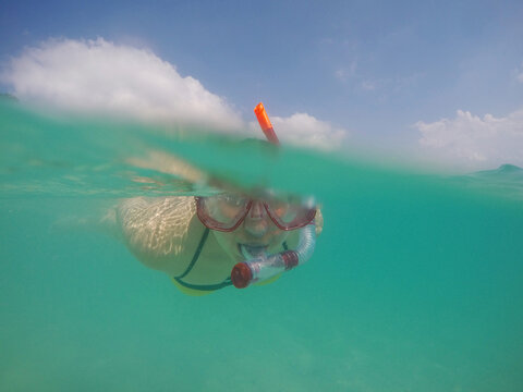 Split-level underwater photo of woman snorkeling in turquoise sea with mask and snorkel, half above water with blue sky and clouds