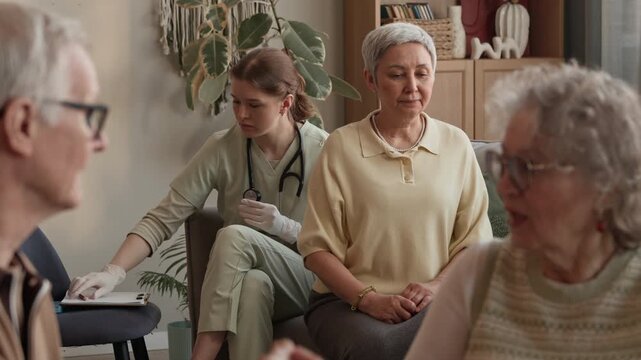 Focus on young Caucasian nurse using stethoscope checking breath of senior woman while elderly couple communicating in foreground in nursing home