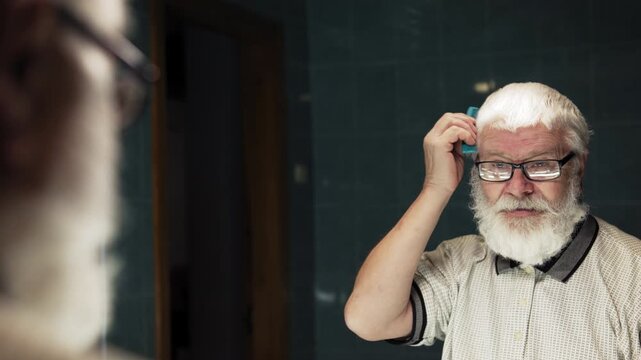 Old man in glasses is combing his beard in bathroom in front of a mirror