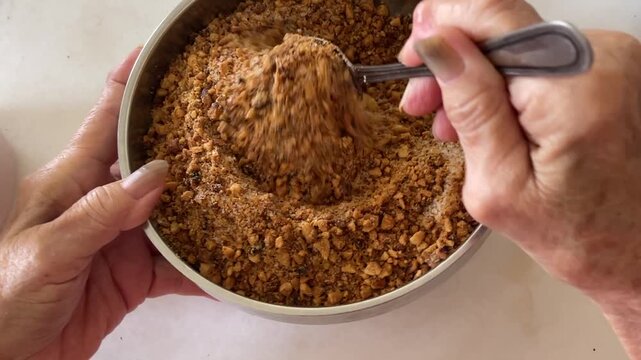 A close-up shot shows hands using a metal spoon to mix coarse roasted peanuts with brown sugar in a bowl, preparing a traditional sweet filling for handmade baozi.