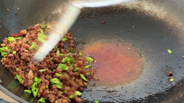 A hand using a spatula to push stir fried minced meat and petai Parkia speciosa beans to the side of a wok to drain excess oil.
