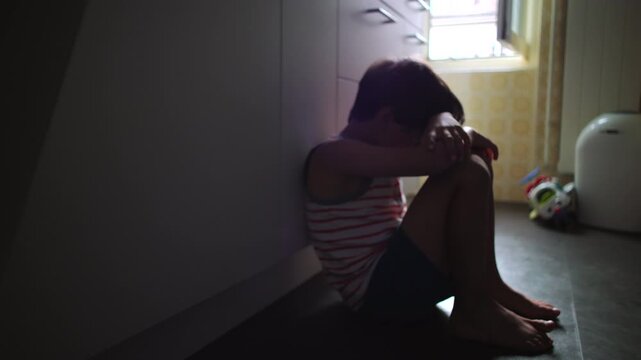 Child sitting alone on kitchen floor near window hugging knees and hiding face expressing deep loneliness depression and childhood trauma in quiet domestic environment suggesting neglect