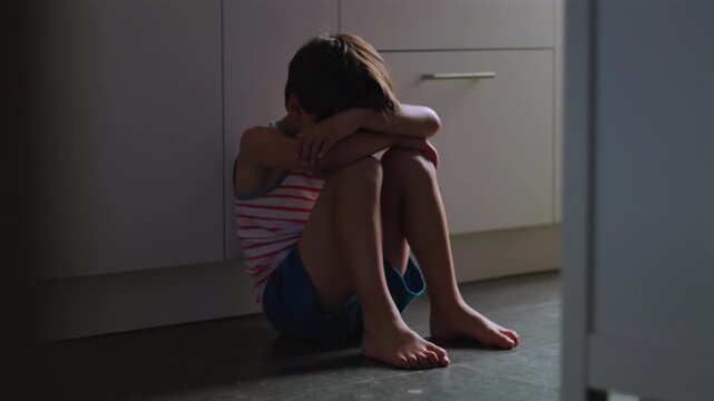 Child sitting alone on kitchen floor with knees drawn close and face hidden in arms expressing deep loneliness depression and emotional trauma in quiet domestic interior