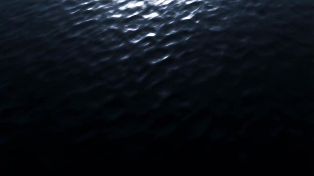 Slow aerial view of ocean surface rippling and reflecting low light across dark open sea expanse at night top down perspective