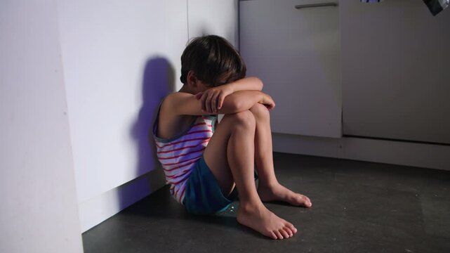 Child sitting on kitchen floor in corner hugging knees and hiding face expressing deep loneliness depression and childhood trauma suggesting emotional neglect and inner suffering at home
