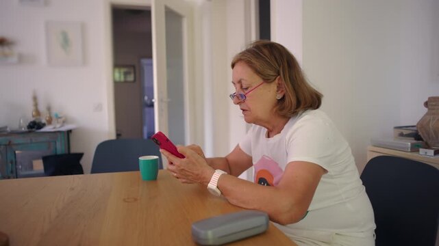 Mature woman sitting at dining table using smartphone, focused expression while reading and typing message, quiet home interior, coffee cup nearby, everyday technology use