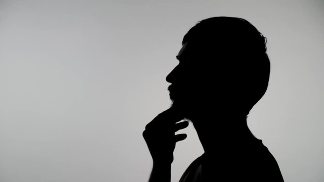 An anonymous silhouette of a man in profile contemplates while scratching his beard against a white wall