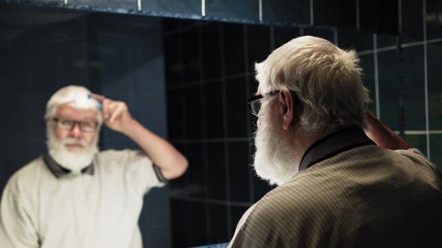 Gray-haired man in glasses is combing his hair in bathroom in front of a mirror
