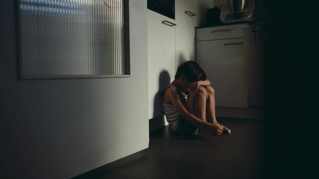 Sad child hiding face while sitting on floor in kitchen corner conveying emotional pain loneliness and fear within domestic space suggesting neglect and inner struggle
