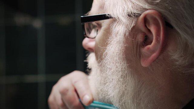 Old man in glasses is combing his white beard in bathroom in front of mirror