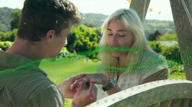 Man proposing wedding, sliding ring onto her finger on bench, green particles sweeping over faces