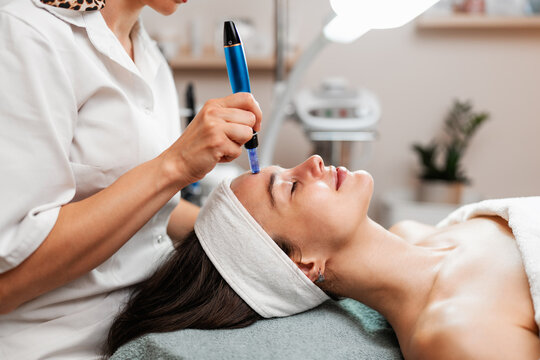 A professional cosmetologist uses a dermapen for a microneedling facial procedure on a young woman lying down in a beauty clinic.