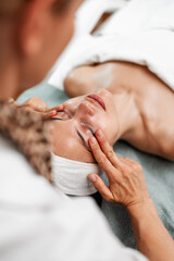 Fototapeta premium A professional aesthetician performs a relaxing facial massage on a young woman in a wellness spa. The client is lying down with eyes closed during the skincare treatment.
