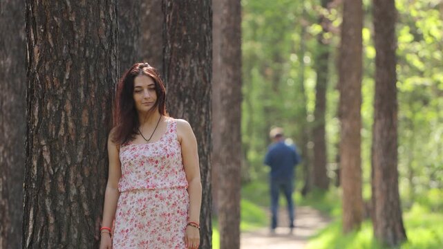 A woman and a man in a forest. A young woman stands, leaning against a pine tree trunk. Behind her on the path is the silhouette of a man.