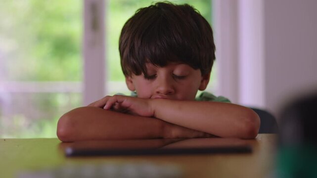 Young boy resting head on folded arms while watching screen on table showing absorbed attention quiet focus and passive entertainment during calm indoor moment at home