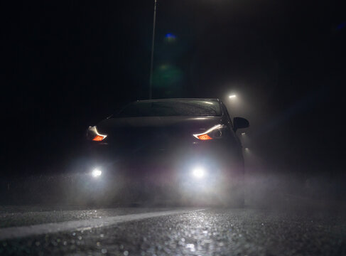 A dark car sits parked on a wet road at night. Its bright headlights, daytime running lights, and fog lights cut through the heavy mist.