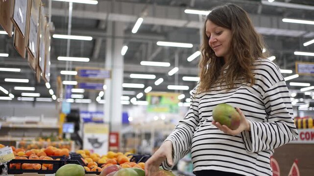 Pregnant young woman choosing fresh organic fruits at the grocery store, carefully selecting and smelling a ripe mango for her healthy diet during her happy pregnancy journey