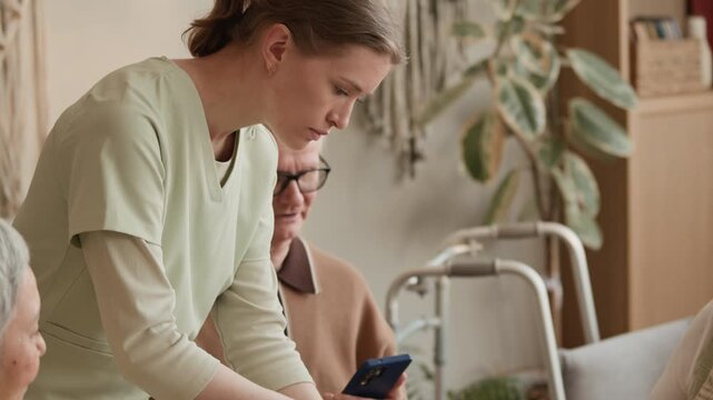 Handheld tilt down shot of young Caucasian nurse cutting cake on table with group of senior people having celebration with tea in care home