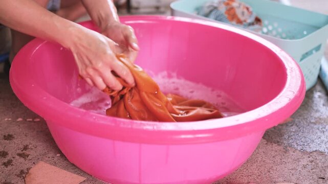 Close-up of hands scrubbing colored clothes in a plastic basin filled with soapy water, representing manual laundry, stain removal, and everyday household cleaning routine.