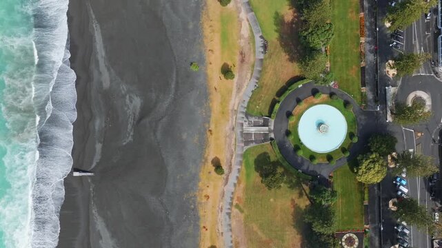 Aerial view of the beach and a circular fountain at Marine Parade, Napier, Hawkes Bay, New Zealand. The turquoise ocean meets the dark sand beach, with a park and street visible.