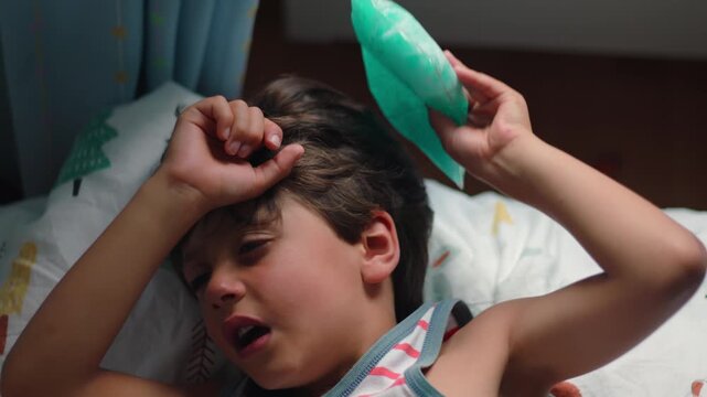 Close view of child applying cold compress to head while lying on pillow expressing pain relief attempt and physical discomfort in home setting