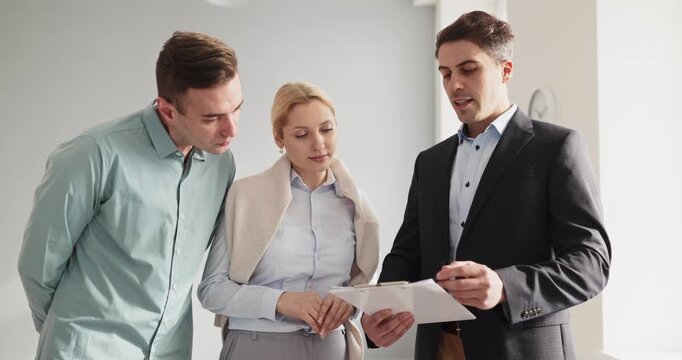 Realtor shows couple apartment during property viewing. Agent outlines options to clients and buyers. Contract terms shown on clipboard in bright interior. Focused real estate sale concept.