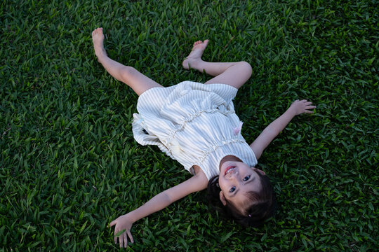 Happy little girl lying on green grass and smiling at camera in a park. Joyful childhood moment, outdoor play, freedom and carefree kid lifestyle enjoying nature and summer day.