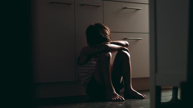 Sad child sitting alone on kitchen floor with arms wrapped around knees hiding face expressing loneliness fear and emotional distress in quiet home interior environment
