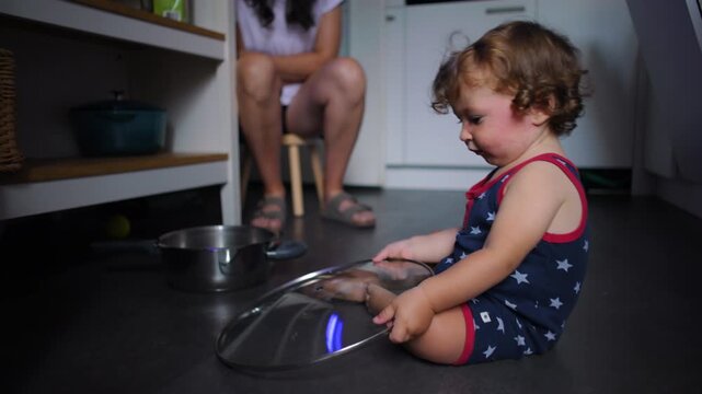 Toddler sitting on kitchen floor holding metal lid and pot, playing with cookware while parent nearby, curious exploration, everyday domestic scene with early learning and focus