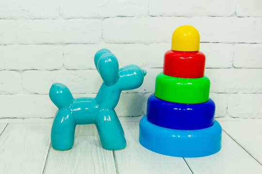 Colorful stacking ring pyramid toy stands beside turquoise balloon dog figurine on white wooden surface against brick wall. Children play, early childhood development, educational toys concept.