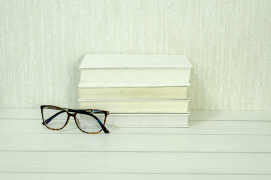 Stack of white books with tortoiseshell eyeglasses on white wooden table. Reading, education, knowledge, study, literature, learning, academic and intellectual concept