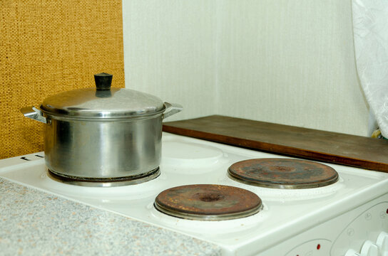 Stainless steel cooking pot with lid on white electric stove featuring worn rusted burners. Domestic kitchen scene illustrating home meal preparation, culinary routine, and household appliance usage.