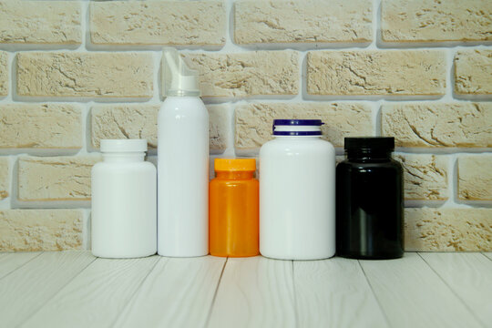 Five medicine bottles and a spray container stand on a white wooden table against a brick wall. Pharmaceutical packaging, healthcare supplies, vitamins, and medical treatment arrangement.