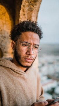 A cinematic close-up photograph of a young man with thick glasses and a look of deep concentration, threading a needle with silver thread by a window, within an old stone archway overlooking a mist...