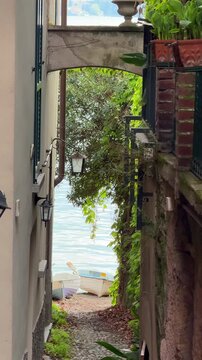 Narrow Italian street leading to Lake Garda with fishing boats on the shore and fresh spring greenery. Peaceful scenic view of lakeside village atmosphere.