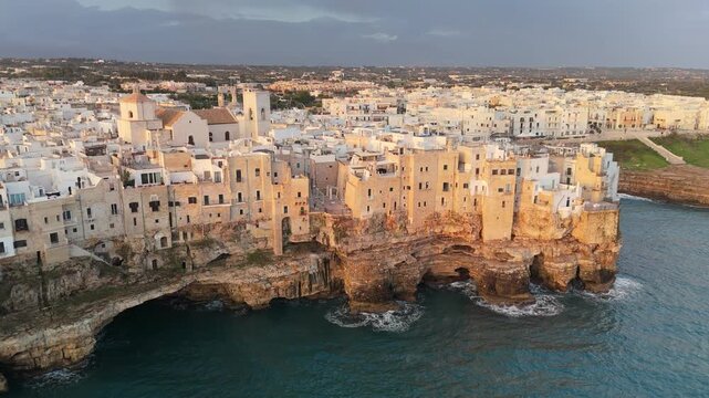 White buildings of the historic center in Polignano a Mare, Italy sit on limestone cliffs. The turquoise Adriatic Sea washes the rocky coast of Polignano a Mare, Italy in this drone aerial