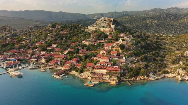 The historic fortress of Simena sits on a peak in the village of Kalekoy, Turkey. Aerial drone shot shows red-roofed houses by the sea in the village of Kalekoy, Turkey